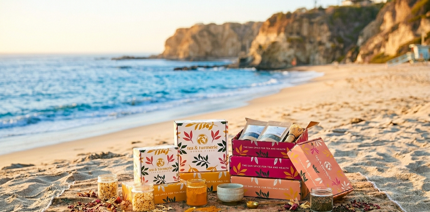 Vibrant branded Tea & Turmeric gift boxes and glass jars of loose-leaf spices arranged on a woven textile over the sand, with the cliffs and ocean of Laguna Beach in the sunny background.