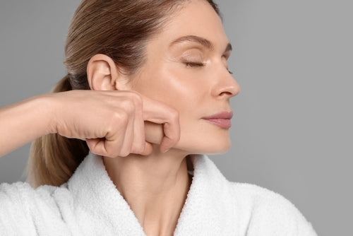Woman massaging her face and jaw in a white robe, practicing Ayurvedic abhyanga self-massage technique