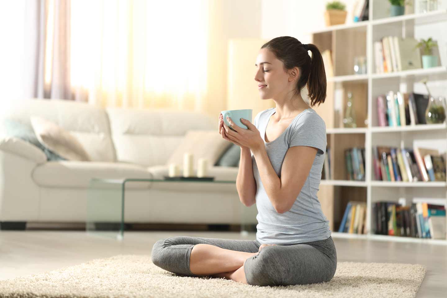Woman sitting cross-legged on floor holding herbal tea cup practicing Pranayama box breathing meditation at home