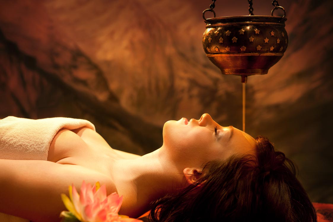 Woman receiving shirodhara, an Ayurvedic oil therapy treatment, lying peacefully as warm oil flows from a traditional copper vessel